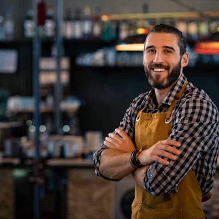 Happy satisfied bartender wearing apron while standing near counter with crossed arms and looking at camera. Confident coffee shop waiter smiling at cafeteria. Successful proud young brewer at his pub with copy space.